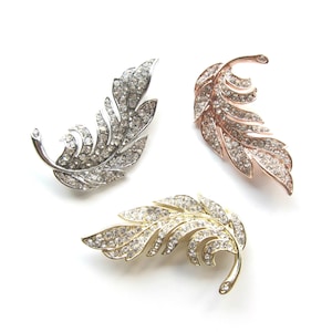 Op de afbeelding: Drie veer-vormige broches met strass. De broches zijn zilver, goud en roségoud.