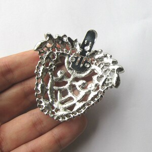 Op de afbeelding: Zilverkleurige metalen broche met een filigraanontwerp en een decoratieve sluiting. De broche heeft de vorm van een hart en heeft een gedetailleerd, ingewikkeld patroon.