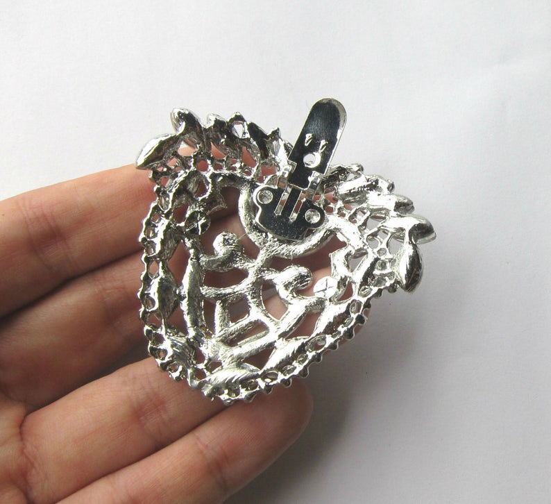 Op de afbeelding: Zilverkleurige metalen broche met een filigraanontwerp en een decoratieve sluiting. De broche heeft de vorm van een hart en heeft een gedetailleerd, ingewikkeld patroon.