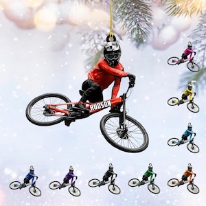 May include: A Christmas ornament featuring a cyclist in a red jacket and helmet on a red bike with the name "HUDSON". Additional ornaments in various colors are also shown. The ornament is suspended by a gold chain.