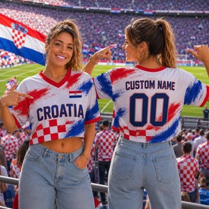 Two women wearing Croatia soccer jerseys with custom names and numbers, standing in front of a stadium filled with spectators.