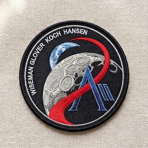 May include: A circular black embroidered patch featuring a stylized moon, Earth, and a red and blue design. The patch has the names "WISEMAN GLOVER KOCH HANSEN" in white lettering around the edge. The patch is likely a commemorative item.
