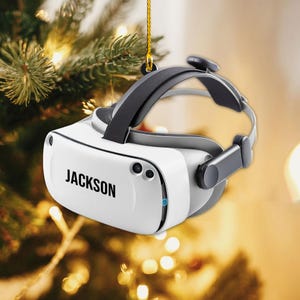 May include: A white VR headset with black accents and the name "JACKSON" printed on the front. The headset is suspended by a gold string, with a blurred background of a Christmas tree.