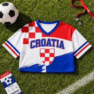 a red, white, and blue Croatia soccer jersey laid out on a grassy surface, along with a soccer ball, a whistle, and a ticket.