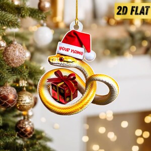 May include: A Christmas ornament featuring a yellow snake wearing a Santa hat with the text "YOUR NAME". The snake is coiled around a red gift box with a red bow. The ornament is 2D flat.