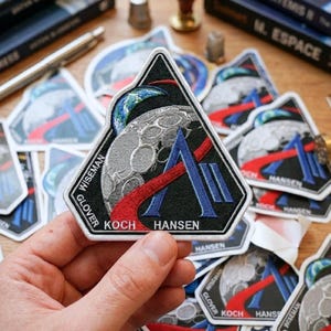 May include: A triangular black patch featuring a stylized moon, Earth, and a red and blue design. The patch has the names WISEMAN, GLOVER, KOCH, and HANSEN. The patch is surrounded by other similar patches.
