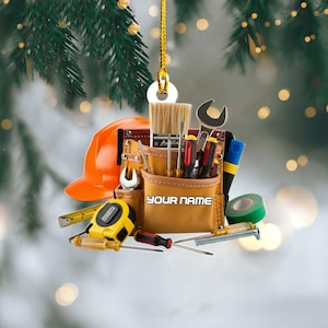 May include: A Christmas ornament featuring a tool belt filled with tools, an orange hard hat, and a yellow tape measure. The text "YOUR NAME" is printed on the tool belt. The ornament is hanging from a gold string.