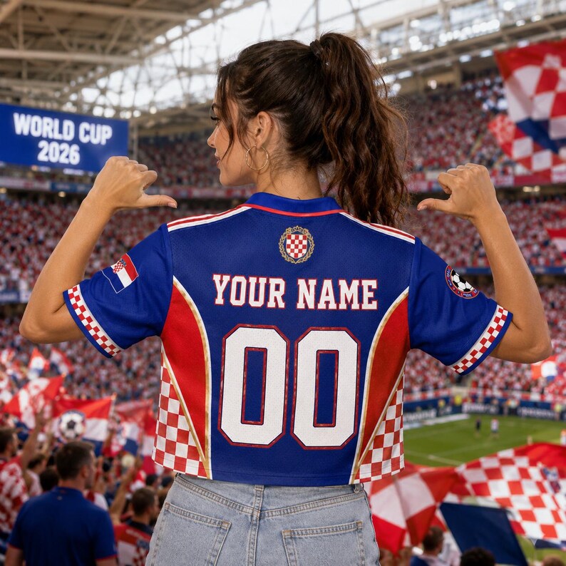 A woman wearing a blue and red soccer jersey with the number 00 and the name "YOUR NAME" printed on the back, standing in a crowded stadium with flags and banners visible in the background.
