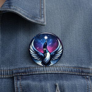 May include: A round button pin featuring a stylized bird with outstretched wings against a starry galaxy background. The bird is primarily black and white with a red eye, reaching towards a bright star. The background has shades of blue, purple, and pink.