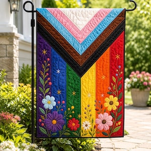 May include: A colorful garden flag featuring a Pride flag design with a chevron pattern and vertical stripes in rainbow colors. The flag is adorned with embroidered flowers and foliage, and is displayed on a black metal stand.