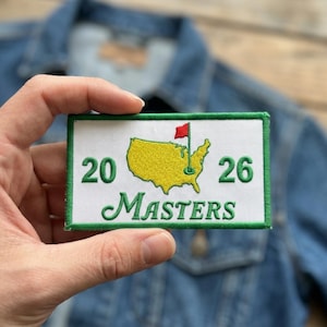 May include: A rectangular patch with a green border and white background. The patch features a yellow map of the United States with a red flag, the number 20 and 26, and the word "MASTERS" in green script.