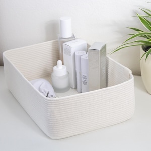 Op de afbeelding: Een rechthoekige, crèmekleurige geweven touwmand gevuld met huidverzorgingsproducten. De mand bevat diverse witte flessen, dozen en een kleine pouch. De producten omvatten een druppelfles, spuitflessen en cilindrische containers.