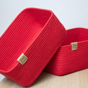 Op de afbeelding: Twee rode rechthoekige opbergmanden van geweven materiaal. Elke mand heeft een klein, rechthoekig label met de woorden "Twist & Tidy" erop gedrukt. De manden staan op een lichtgekleurd houten oppervlak.