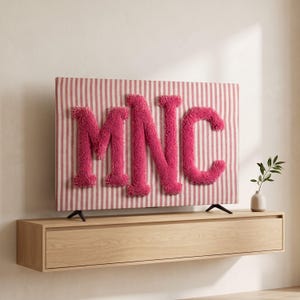 Könnte beinhalten: Eine rechteckige Fernsehbildschirmabdeckung mit einem vertikalen Streifenmuster in Rosa und Weiß. In der Mitte befinden sich die Buchstaben MNC als große, erhabene, strukturierte, pinkfarbene getuftete Garnbuchstaben mit weicher Optik.