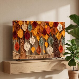 May include: A television screen displaying a digital artwork of a forest scene featuring numerous hanging macrame-style leaf ornaments in shades of yellow, orange, red, brown, cream, and sage green, with tree trunks and a woven rope border.