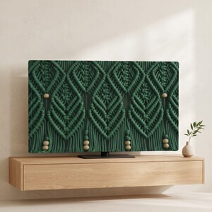 Pode incluir: Uma tela de televisão exibindo uma tapeçaria de macramé verde-escura com um padrão de diamante repetitivo e contas de madeira clara integradas no design de corda atada, apresentando elementos de franja verticais na borda inferior.