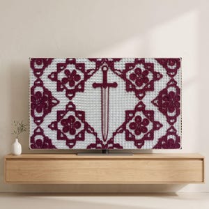 May include: A television screen displaying a white fabric background with a repeating dark red crocheted floral pattern and a central dark red crocheted dagger motif. The screen is mounted on a light wood floating shelf against a plain wall.