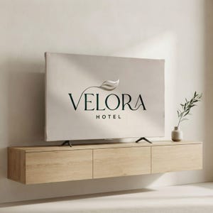 Puede incluir: Un televisor de pantalla plana que muestra el texto VELORA HOTEL con un gráfico de hoja sobre la palabra VELORA, colocado sobre un mueble multimedia flotante de madera clara con tres cajones, junto a un pequeño jarrón con una rama verde.