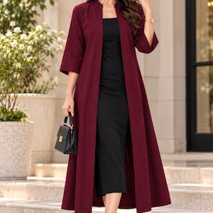 Peut inclure: Un long manteau duster bordeaux ouvert sur le devant avec des manches trois-quarts, porté sur une robe midi noire simple. La tenue est assortie d'un petit sac à main noir structuré et de sandales à brides noires, formant un ensemble superposé.