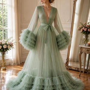 May include: A floor-length sage green gown made of sheer tulle fabric. The dress features a deep V-neckline, long flowing sleeves with tiered ruffled cuffs, and a matching tie-waist belt. The skirt is voluminous with multiple layers of ruffles.