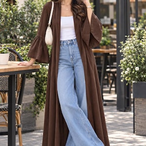 Puede incluir: Un cárdigan largo de color marrón oscuro abierto con mangas acampanadas, usado sobre una camiseta sin mangas blanca y jeans anchos de mezclilla azul claro. El conjunto se combina con zapatillas blancas y un pequeño bolso de hombro color crema.
