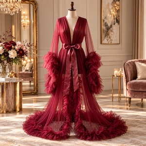 May include: A floor-length burgundy robe made of sheer pleated tulle fabric. The garment features long sleeves with voluminous ruffled cuffs, a deep V-neckline, and a matching satin ribbon tie at the waist. The hem is finished with a wide ruffle.