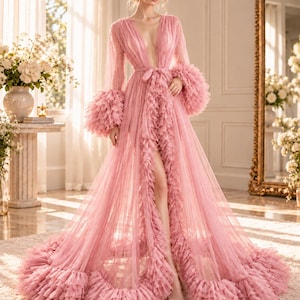 May include: A floor-length dusty pink robe made of sheer tulle fabric. The garment features a deep V-neckline, long sleeves with voluminous ruffled cuffs, and a matching tie belt at the waist. The front opening and hem are trimmed with layers of ruffles.