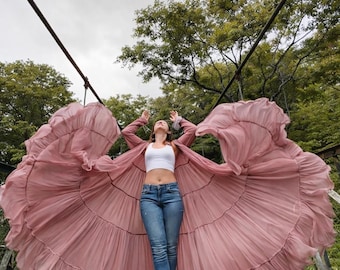 May include: A person stands on a bridge, wearing a white crop top, blue jeans, and a long, flowing, dusty rose-colored duster. The duster is open, creating a wide, circular shape. The background features lush green trees and a cloudy sky.