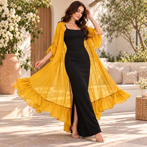 Puede incluir: Un vestido negro largo hasta el suelo con tirantes finos y una abertura lateral, combinado con un cárdigan largo y transparente de color amarillo mostaza con mangas tres cuartos y un dobladillo con volantes que se abre.