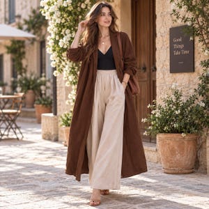 May include: A long-sleeved brown duster coat worn over a black V-neck camisole and high-waisted beige wide-leg trousers. The outfit is paired with brown open-toe sandals and a brown shoulder bag. The ensemble features a clean, layered aesthetic.
