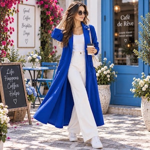 Pode incluir: Um casaco comprido azul vibrante usado sobre uma camisola sem mangas branca e calças brancas de perna larga. O conjunto é combinado com ténis brancos e uma pequena mala de ombro bege. O casaco tem mangas compridas e dois bolsos frontais.