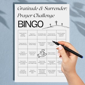 May include: A white Bingo card with the text "Gratitude & Surrender: Prayer Challenge" and "BINGO". The card has a grid of squares with prompts like "Thank God for today's blessings" and "Pray for a grateful heart". A hand holds a pen.