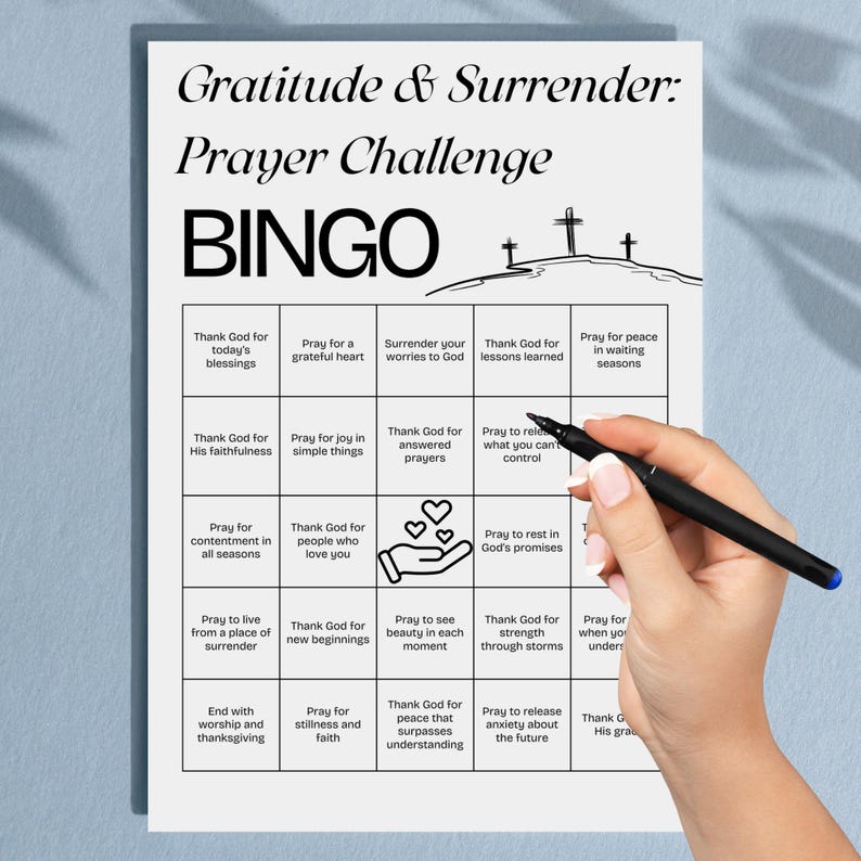 May include: A white Bingo card with the text "Gratitude & Surrender: Prayer Challenge" and "BINGO". The card has a grid of squares with prompts like "Thank God for today's blessings" and "Pray for a grateful heart". A hand holds a pen.