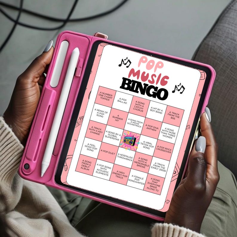 May include: A pink tablet with a Pop Music Bingo game displayed on the screen. The game board features various music-related prompts. A pink stylus is attached to the tablet case. The tablet is held by a person.