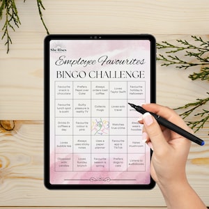 May include: A digital tablet shows an "Employee Favourites BINGO CHALLENGE" game. The game board includes prompts such as "Loves Taylor Swift" and "Hates mushroom." A hand with a pen is positioned over the screen.