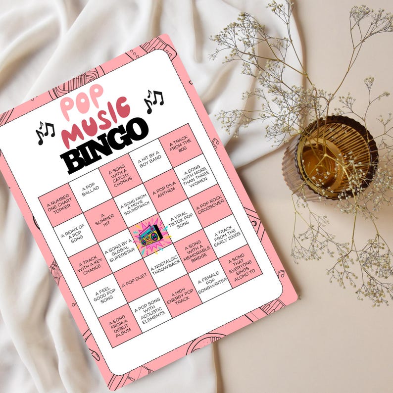 May include: A pink Pop Music Bingo game card with black text and musical note graphics. The card features a grid of bingo squares with various pop music-related prompts. A decorative vase with flowers is in the background.