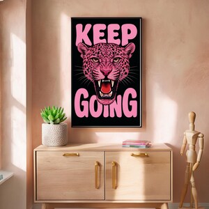 Puede incluir: Una impresión artística enmarcada con una ilustración de leopardo rosa y las palabras "KEEP GOING" en rosa. La obra de arte está sobre un fondo negro. La impresión se exhibe en un mueble de madera con una planta suculenta en una maceta blanca.