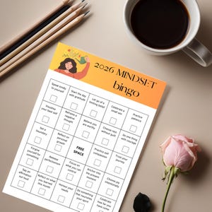 Könnte beinhalten: Eine Mindset-Bingo-Karte für 2026 mit einer floralen Illustration und dem Titel "2026 MINDSET bingo". Die Karte enthält Aufforderungen zur Selbstverbesserung, wie "Sprich freundlich zu dir selbst" und "Feiere einen kleinen Erfolg". Eine Tasse Kaffee, Bleistifte und eine Rose sind ebenfalls im Bild.