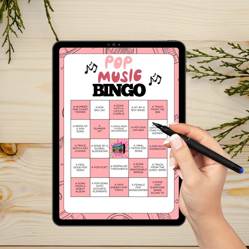 May include: A tablet displays a pink Pop Music Bingo game. The game board features various music-related prompts, including "A Song with a Catchy Chorus" and "A Viral TikTok Pop Song." A hand holding a pen points to the board.