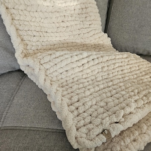 Puede incluir: Una manta de punto grueso de color crema doblada sobre un sofá gris. La manta tiene un aspecto grueso y texturizado, con bucles visibles y una sensación suave y afelpada. Los bordes están cuidadosamente acabados y se adjunta una pequeña etiqueta. La manta parece acogedora y acogedora.