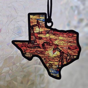 May include: A Texas-shaped stained glass ornament with a black border. The interior features a mosaic design with oil derricks, and a color palette of red, orange, yellow, and blue. A black cord is attached for hanging.