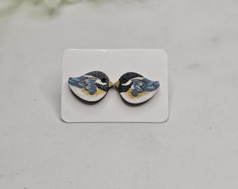 May include: A pair of wooden bird-shaped earrings. Each earring features a black head, blue and white wings, and a yellow beak. The earrings are on a white card, perfect for gifting or display.