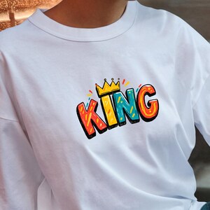 May include: A white short-sleeved t-shirt featuring a graphic print on the chest. The design displays the word KING in bold, colorful, cartoon-style letters with a yellow crown icon positioned directly above the letter I in the center.