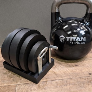 May include: A black kettlebell and a set of black weight plates on a black stand. The kettlebell has a silver handle and the words "TITAN FITNESS" printed on it. The weight plates are stacked and ready for use.