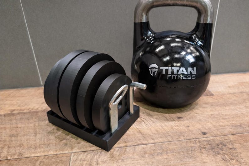May include: A black kettlebell and a set of black weight plates on a black stand. The kettlebell has a silver handle and the words "TITAN FITNESS" printed on it. The weight plates are stacked and ready for use.