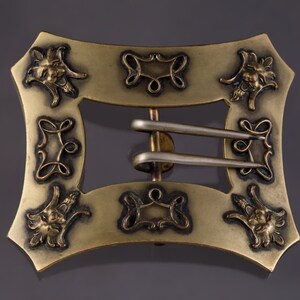 May include: A gold-toned metal buckle with a decorative design featuring stylized faces and swirls. The buckle has a silver-toned metal clasp.