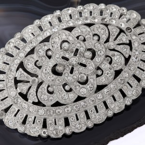 K&ouml;nnte beinhalten: Eine ovale Brosche aus Silber mit einem filigranen Design aus klaren Strasssteinen. Die Brosche ist in einen silbernen Metallrahmen gefasst und hat ein zartes Blumenmuster.