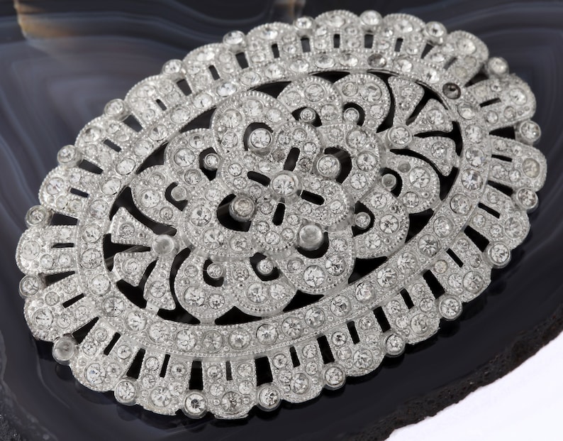 K&ouml;nnte beinhalten: Eine ovale Brosche aus Silber mit einem filigranen Design aus klaren Strasssteinen. Die Brosche ist in einen silbernen Metallrahmen gefasst und hat ein zartes Blumenmuster.