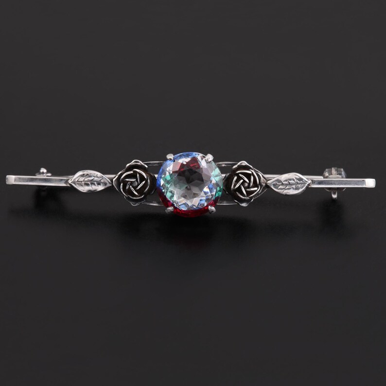 May include: A silver brooch with a large, faceted, clear gemstone surrounded by two silver rose motifs. The brooch has a delicate, vintage style.
