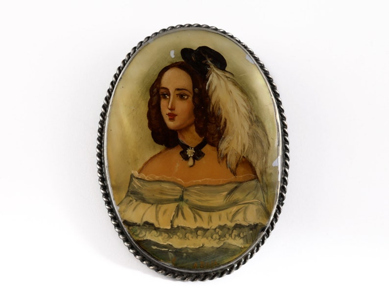 Puede incluir: Broche ovalado antiguo con un retrato pintado a mano de una mujer. El retrato presenta a una mujer con cabello casta&ntilde;o, que lleva un sombrero negro con una pluma y un vestido blanco. El broche tiene un borde plateado retorcido.